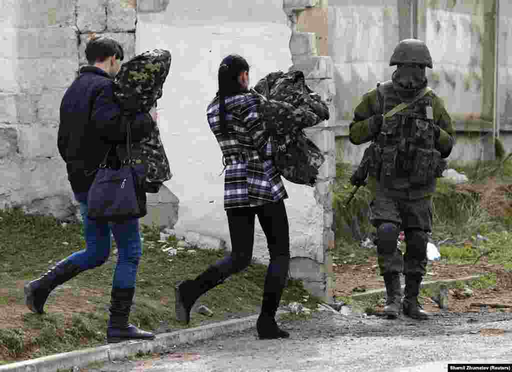 Две женщины, предположительно украинские военные, неся в руках военную форму, покидают военную базу и проходят мимо российского солдата, 19 марта 2014 года.  