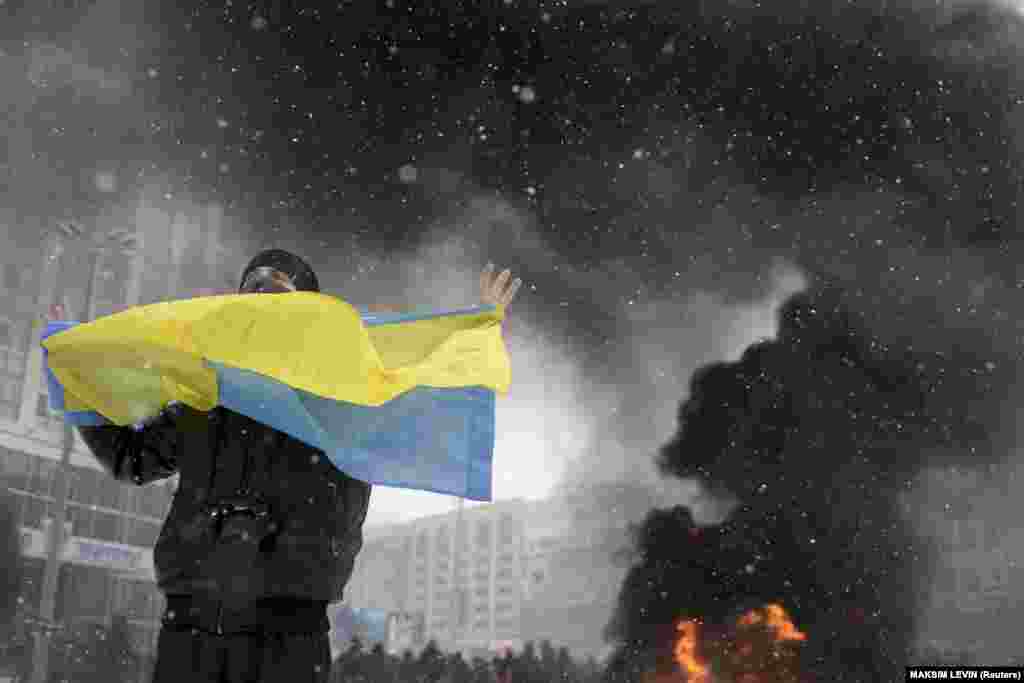 Мужчина держит украинский флаг на фоне столкновений между спецназом и протестующими во время «Революции достоинства» в Киеве, фото от 22 января 2014 года