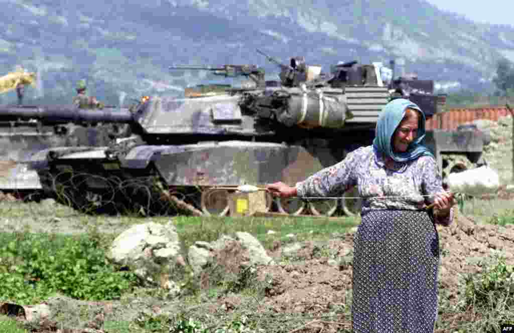 Албанська жінка пряде вовну перед американським танком M1 Abrams, який був частиною операції НАТО під час конфлікту в Косові, біля авіабази Рінас,12 травня 1999 року. З екіпажем із чотирьох осіб (командир, навідник, заряджальник і водій) «Абрамс» є одним із найважчих танків на озброєнні, вагою в майже 62 метричні тонни  