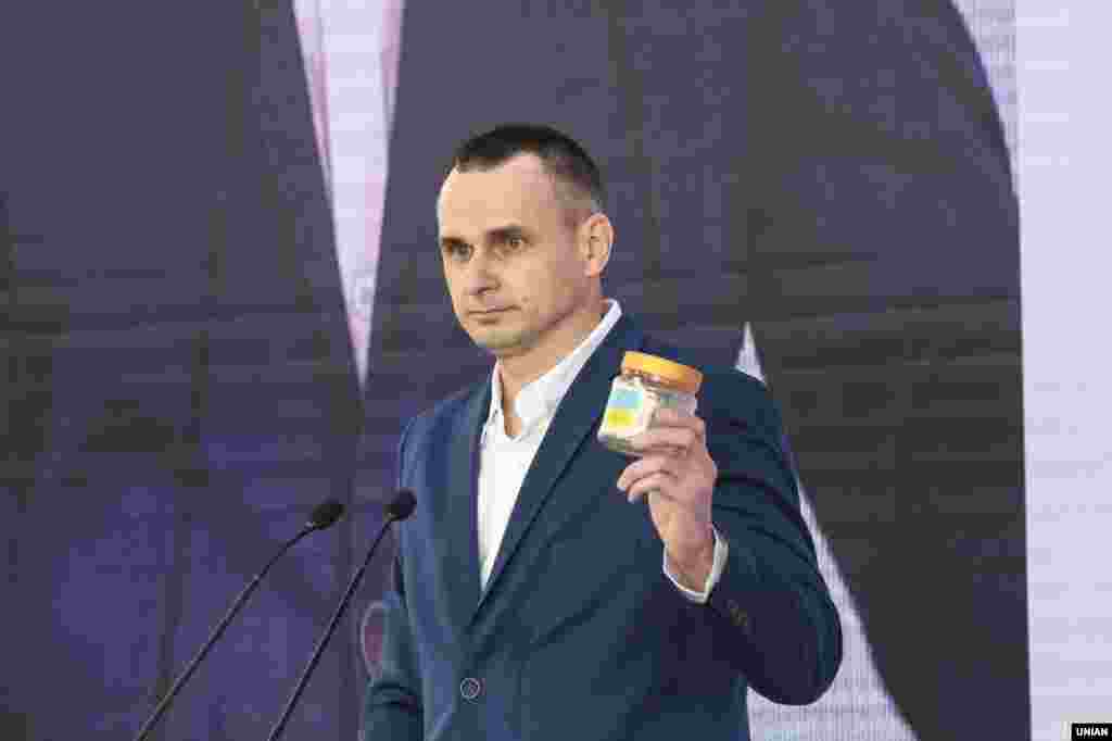 Олег Сенцов во время выступления на 16-м форуме «Ялтинской европейской стратегии», в Киеве, 13 сентября 2019 года. Режиссер рассказал, что после освобождения забрал с собой на память пластиковую банку, в которой он хранил чай в местах лишения свободы в России, и на которую наклеил две полоски в виде украинского флага, и написал фразу «Слава Украине». Эта банка была с ним все пять лет пребывания в неволе. При этом он отметил, что в тюрьмах России у заключенных есть так называемые нагрудные знаки, и свой знак с красной полоской, которая означает «склонен к побегу», он сохранил. В связи с этим он положил свой нагрудный знак в банку и призвал президента Владимира Зеленского, чтобы она была заполнена нагрудными знаками других украинских политузников после их освобождения из российского заключения