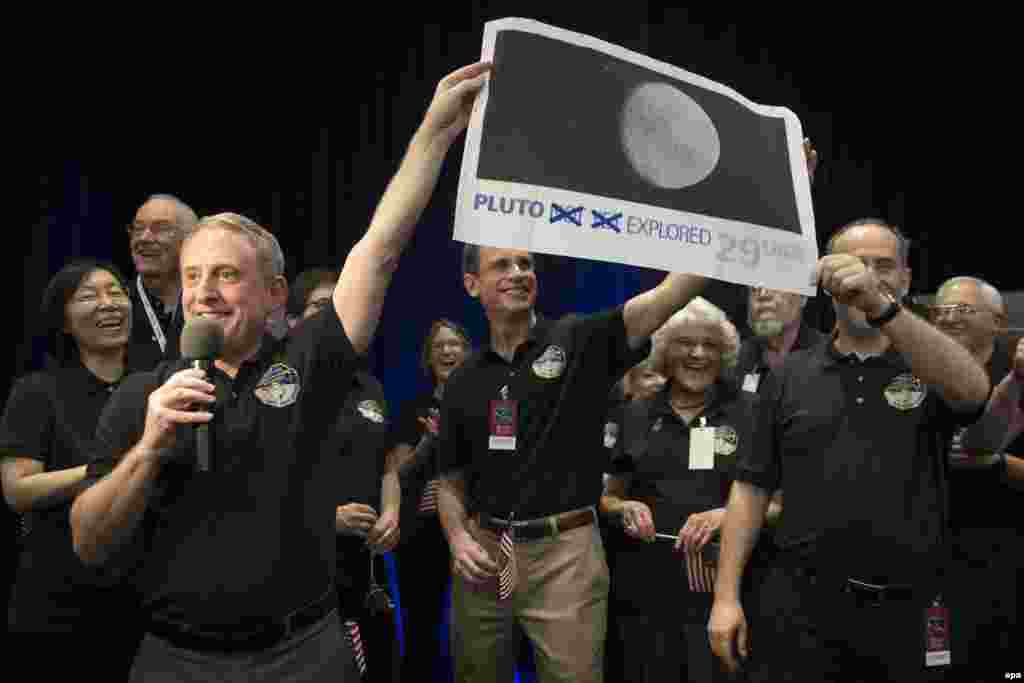 Ученые в центре контроля миссии New Horizons с увеличенным изображением 29-центовой почтовой марки, теперь устаревшей: на ней написано "Плутон еще не исследован" - на изображении слова "еще не" зачеркнуты. 